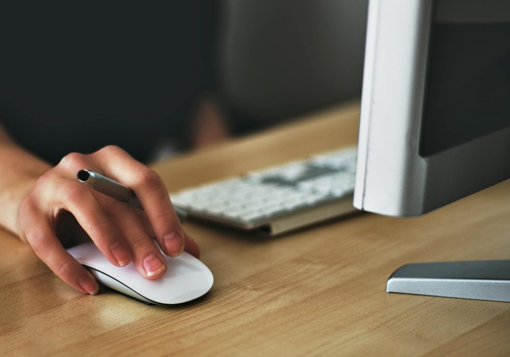 A hand using a wireless mouse at a modern desk setup with a computer and keyboard.